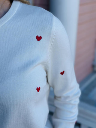 Cream sweater with small red embroidered hearts hanging on a black hanger

Val Sweater featuring red heart embroidery on a soft ivory knit

Heart-embroidered cream sweater displayed against patterned wallpaper

Women’s ivory knit sweater with scattered red heart details