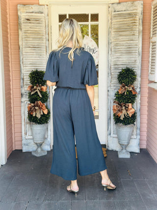 Woman wearing Annalise Evergreen Jumpsuit with ruffle collar and waist tie

Back view of Annalise Evergreen Jumpsuit with keyhole button closure

Close-up of V-neckline and ruffle collar on Annalise Evergreen Jumpsuit