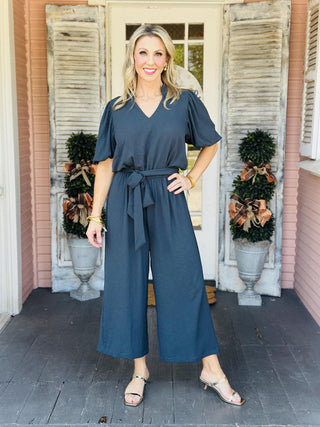 Woman wearing Annalise Evergreen Jumpsuit with ruffle collar and waist tie

Back view of Annalise Evergreen Jumpsuit with keyhole button closure

Close-up of V-neckline and ruffle collar on Annalise Evergreen Jumpsuit