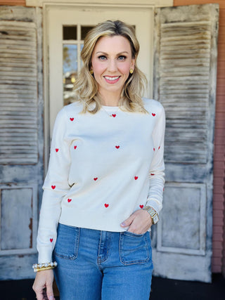 Cream sweater with small red embroidered hearts hanging on a black hanger

Val Sweater featuring red heart embroidery on a soft ivory knit

Heart-embroidered cream sweater displayed against patterned wallpaper

Women’s ivory knit sweater with scattered red heart details