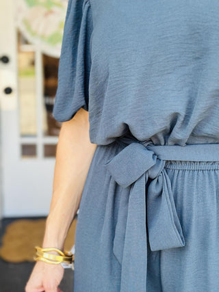 Woman wearing Annalise Evergreen Jumpsuit with ruffle collar and waist tie

Back view of Annalise Evergreen Jumpsuit with keyhole button closure

Close-up of V-neckline and ruffle collar on Annalise Evergreen Jumpsuit