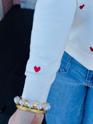 Cream sweater with small red embroidered hearts hanging on a black hanger

Val Sweater featuring red heart embroidery on a soft ivory knit

Heart-embroidered cream sweater displayed against patterned wallpaper

Women’s ivory knit sweater with scattered red heart details