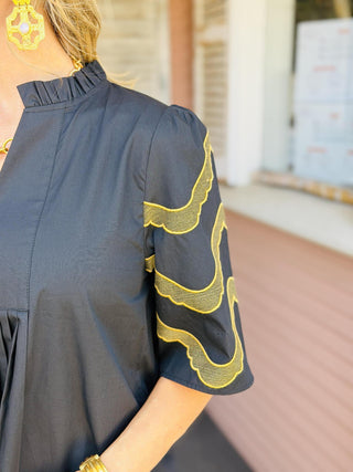 Florence Dress in black featuring embroidered short sleeves, front pleat detailing, a ruffled neckline, and side pockets.