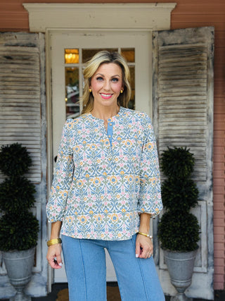 Riley Ashdown Top featuring a cream background with blue, green, blush pink, and golden floral print, split neckline with contrast trim, decorative button detail, and three-quarter puff sleeves.